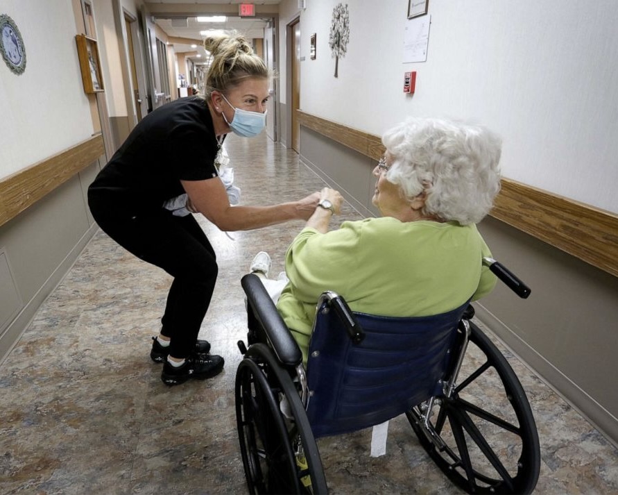 Photo of an elderly person in a wheelchair with a nurse bending down and holding a hand with eachother